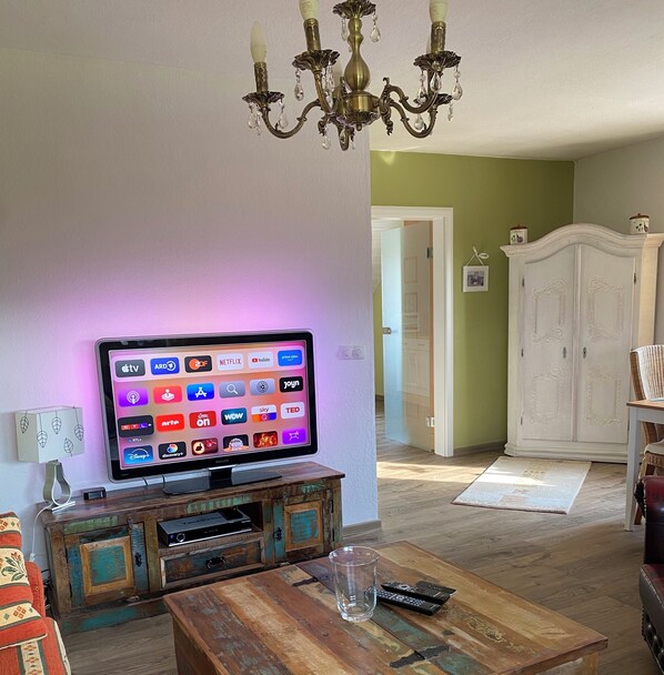 Un salon avec un téléviseur à écran plat fixé à un meuble en bois, un lustre et une armoire blanche en arrière-plan.