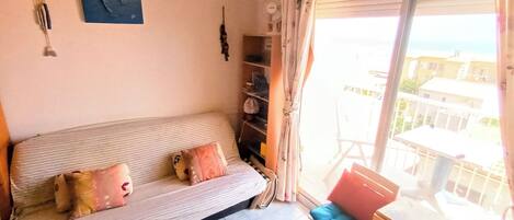 Uma sala de estar com sofá, uma poltrona, uma mesa pequena, janela com cortinas e vista para edifícios.