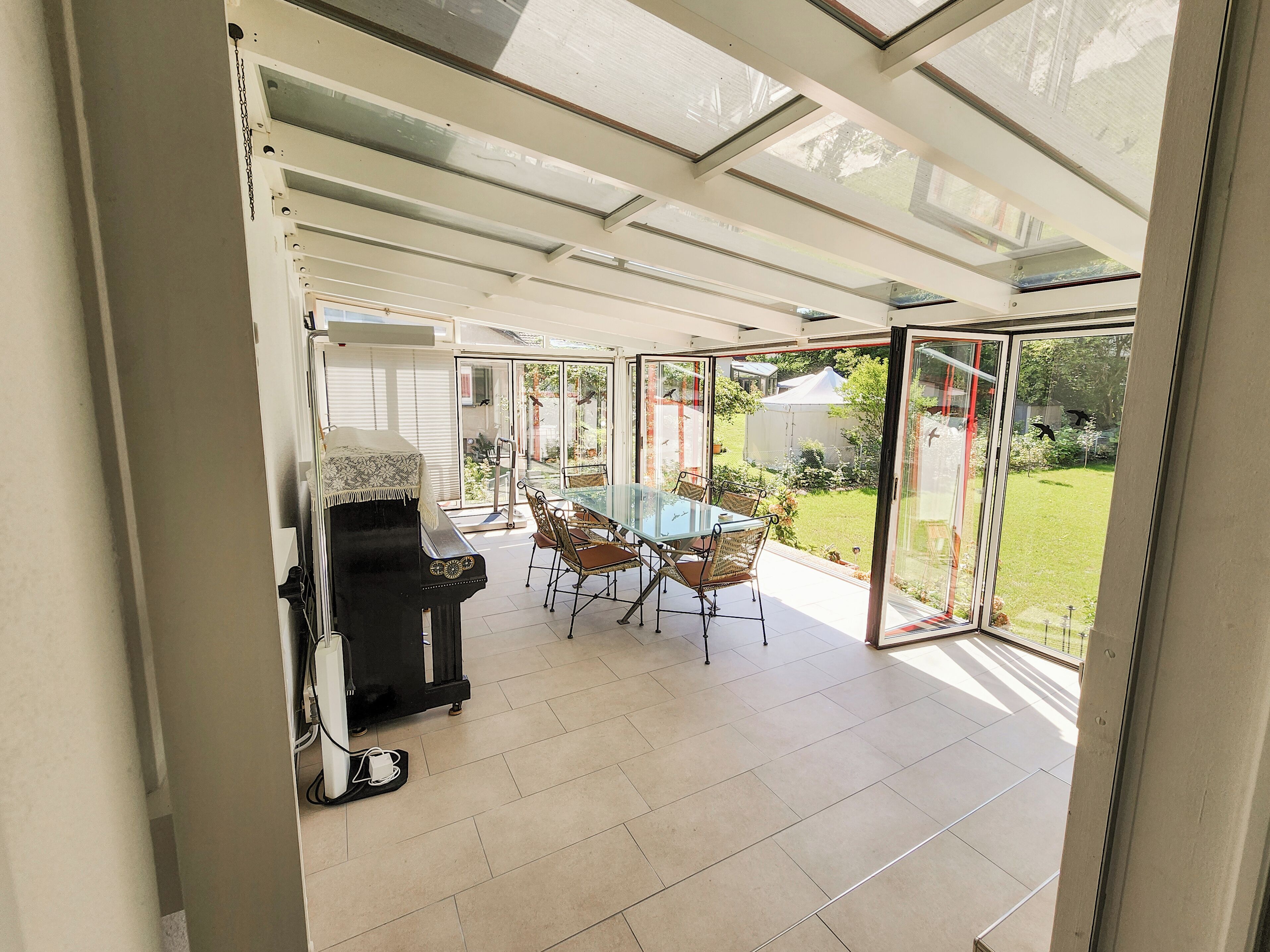Une véranda dotée d’une porte vitrée donnant sur un patio. À l’intérieur, on trouve une table en verre entourée de chaises et un barbecue noir.