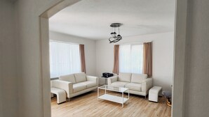 Una sala de estar moderna con suelo de madera, dos sofás blancos, una mesa de centro de vidrio y una lámpara en el techo.