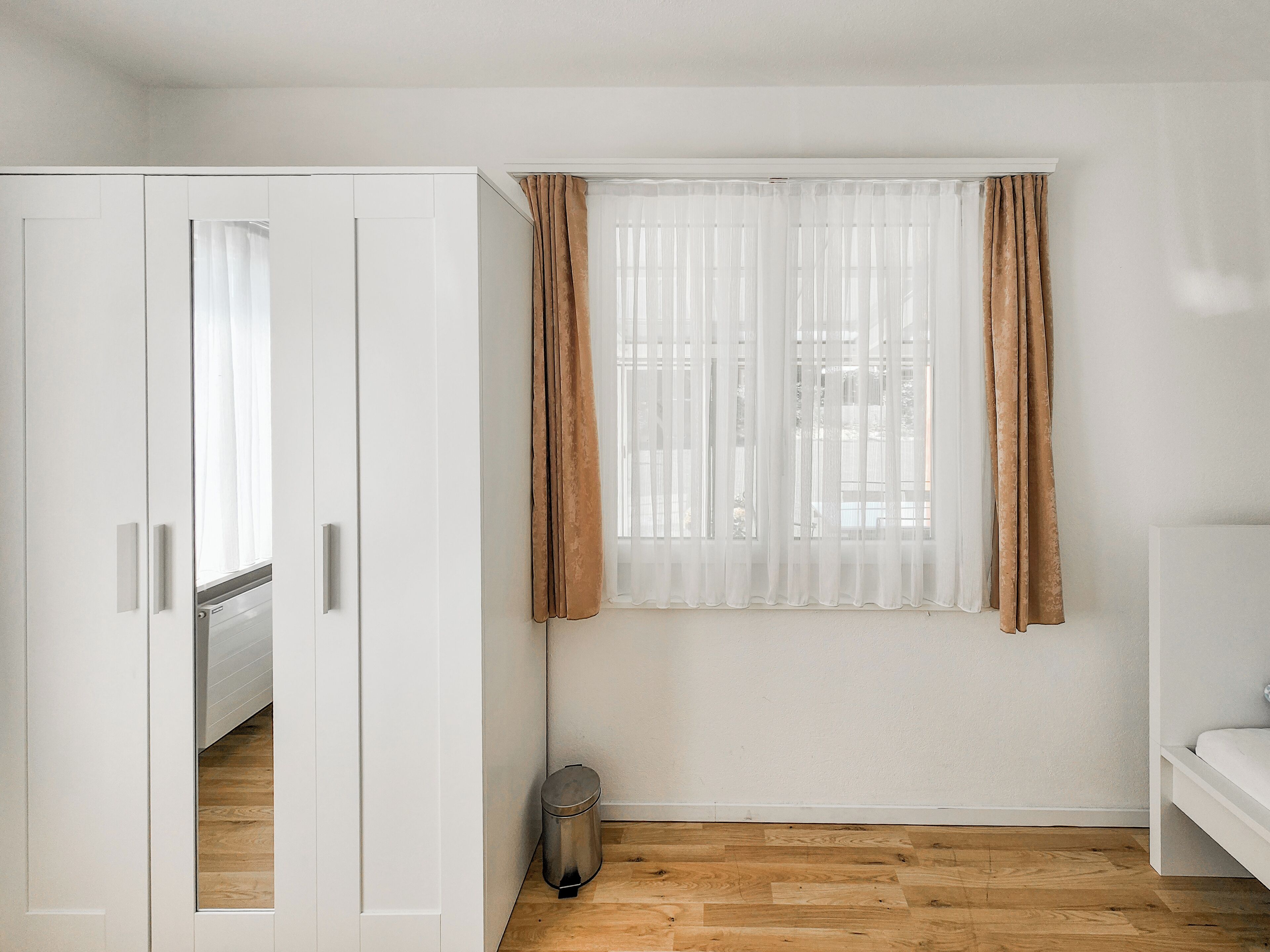 Una habitación con piso de madera, un armario blanco, una ventana con cortinas de tela blanca y cortinas marrones, y un cesto de basura pequeño.
