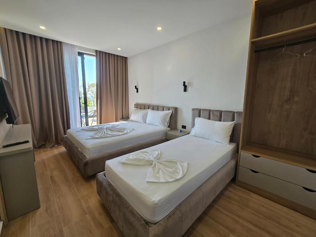 Habitació d'hotel amb dos llits, un armari de fusta, una taula de treball i una finestra gran amb cortines.