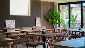 Un restaurant moderne avec des tables en bois et des chaises en osier, de grandes fenêtres et des plantes d’intérieur.