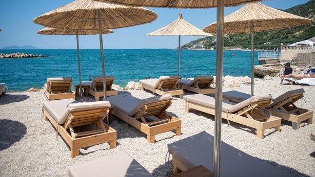 Une plage avec des chaises longues en bois et des parasols en paille, un rivage rocheux et une vue dégagée sur la mer.