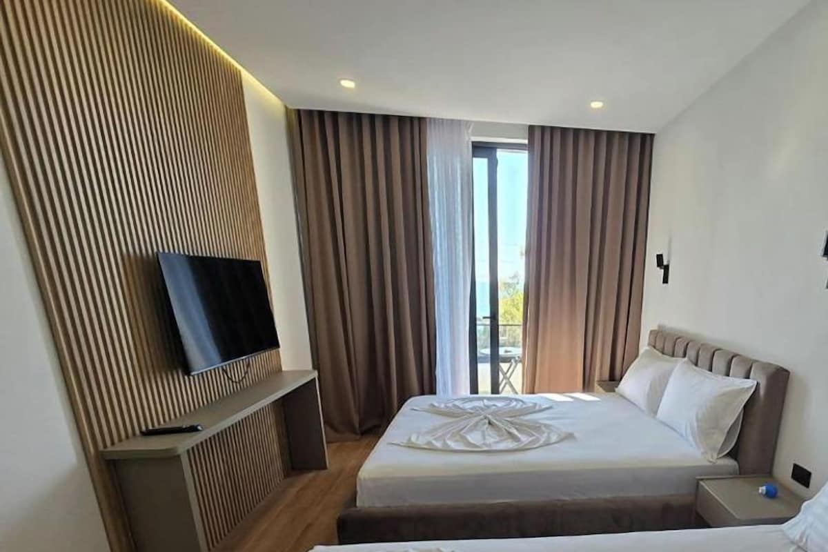 Habitación de hotel con dos camas, un televisor sobre un panel de madera, un balcón con cortinas y vistas al exterior.