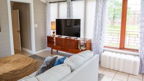 A living room with a sofa, a TV on a wooden cabinet, a patterned rug, and a view through a window with curtains. - Game Room/Foosball/3BD/Families/Coffee Bar/Poker (Des Moines)