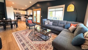 A modern living room with a grey sofa, a coffee table, and a patterned rug. There is a dining area with a wooden table and chairs, and a kitchen in the background. - Entire rental unit. 5 guests. Large private deck. 2 beds. 1 sofa bed. 1 bath. (Belgrade)