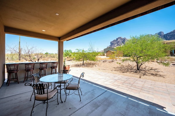 An outdoor patio with a round table and chairs, a view of a desert landscape, and a mountain in the background.