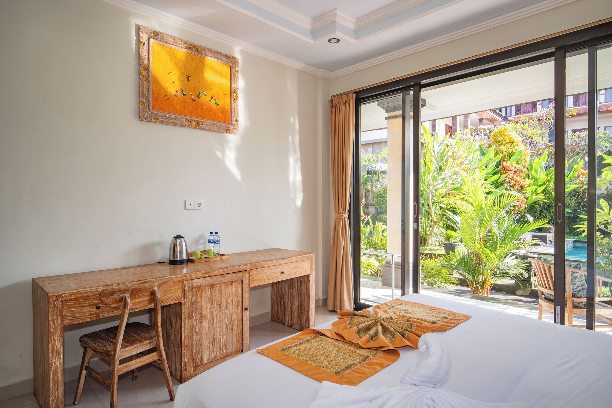 Habitación de hotel con un escritorio de madera, una cama con cubrecama blanco, un ventanal con vista a un jardín y piscina, y un cuadro en la pared.