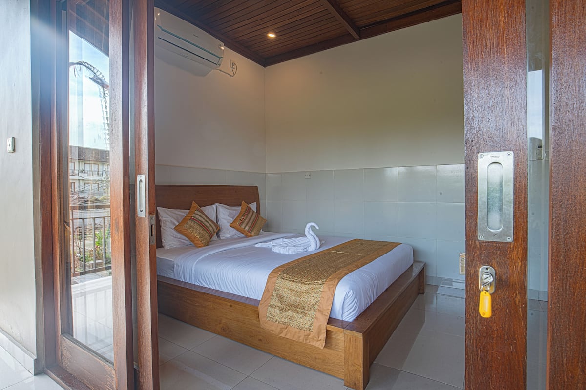 A wooden bed with white bedding and a decorative swan pillow, a wooden door with a metal handle, and a view of the outside balcony.