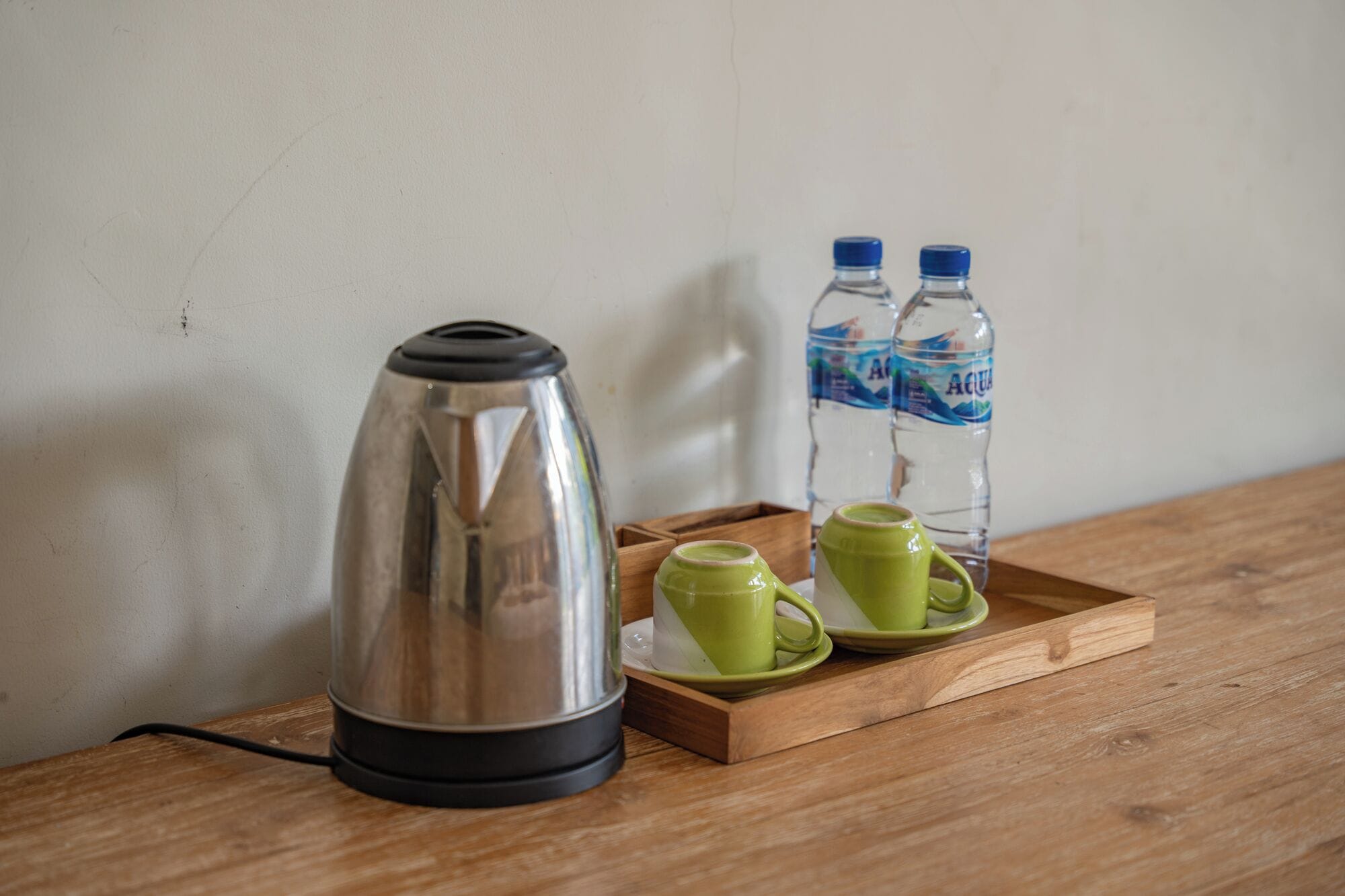 Una tetera eléctrica de acero inoxidable, dos tazas verdes y dos botellas de agua sobre una bandeja de madera.