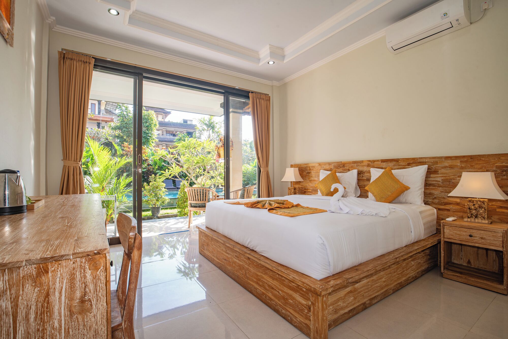A spacious bedroom with a large bed, a wooden headboard, a nightstand, and a view of a lush garden through a sliding glass door.