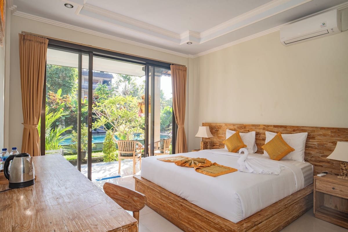 A hotel room with a large bed, a wooden headboard, a desk, and a view of a pool and greenery outside.