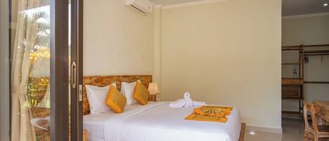 A hotel room with a wooden headboard, a bed with white bedding and orange pillows, a white towel, and a wooden side table with a lamp.