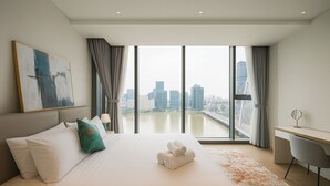 Ein modernes Hotelzimmer mit einem großen Bett, einem Schreibtisch und Blick auf eine Stadtkulisse durch raumhohe Fenster.