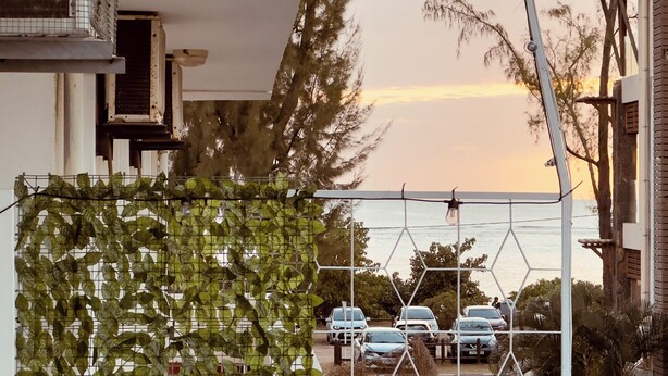 A modern building with a white exterior, a balcony with a metal railing, and a view of parked cars and trees in the background. - Kazafibi B4 - Steps away from the beach (Flic en Flac)