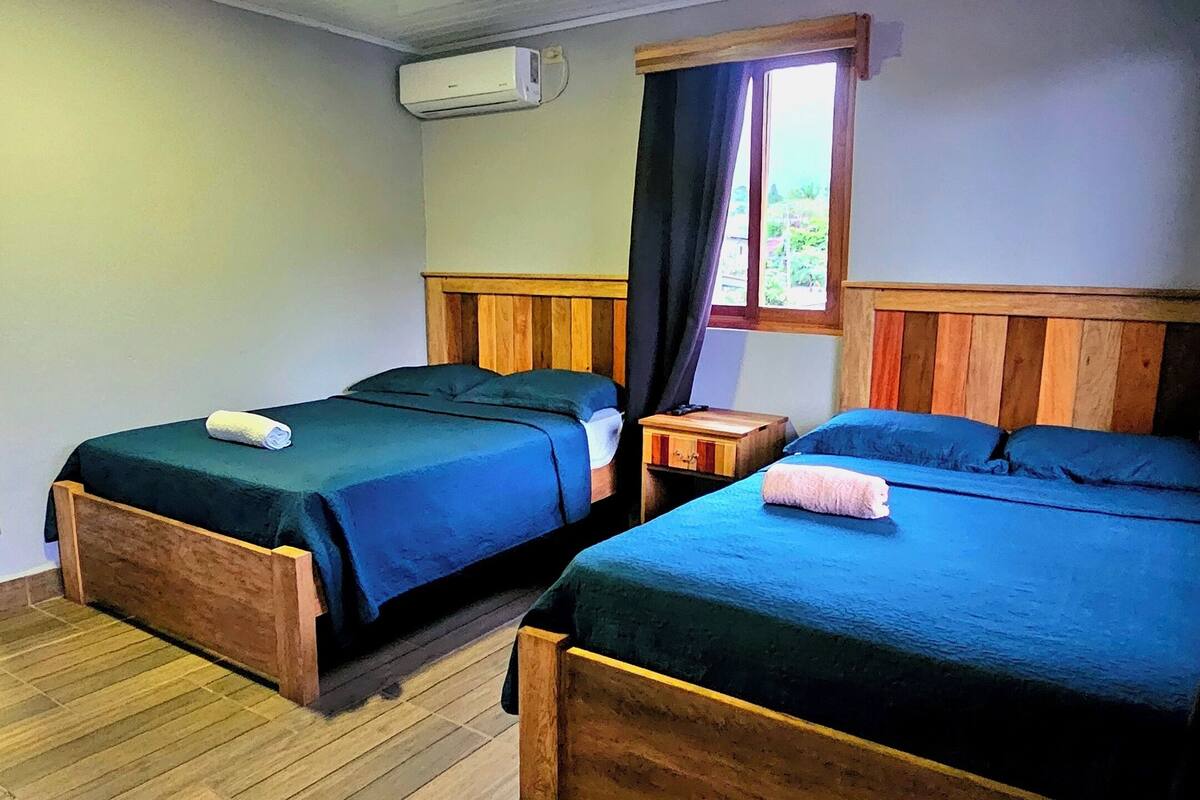 Une pièce avec deux lits en bois, une fenêtre avec des rideaux, un plancher en bois et un appareil de climatisation fixé au mur.
