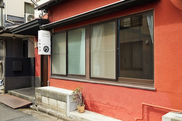A building with a red exterior, large windows, and a small potted plant on the ground. - Nisshin Geppo (Kawasaki)