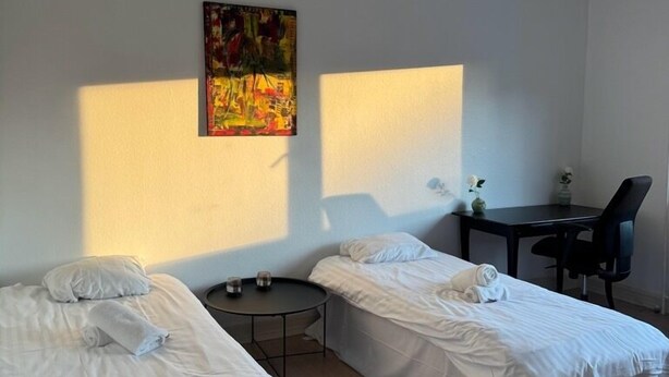 A room with two single beds, a small round table, a desk with a chair, and a painting on the wall.