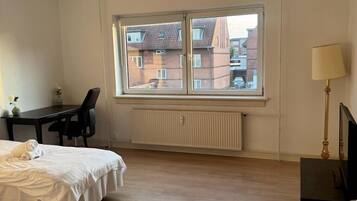 A bedroom with a bed, a desk, a chair, a radiator, and a window with a view of buildings.