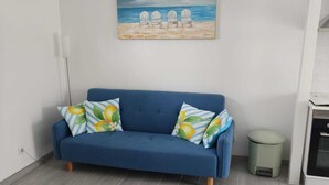 Un canapé bleu avec des coussins à motifs floraux, un tableau représentant des chaises de plage et une poubelle.