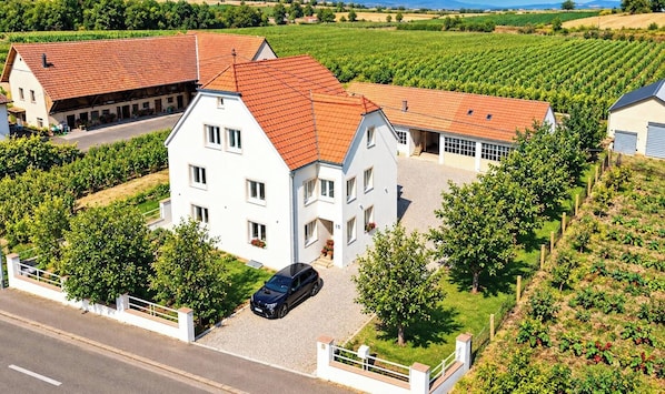 A two-story house with a red-tiled roof, surrounded by vineyards, and a black car parked in the driveway.
