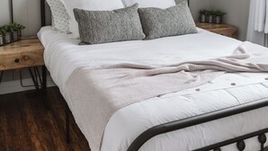 Una cama bien hecha con estructura metálica, ropa blanca y varios almohadones. Hay dos mesitas de noche con lámparas y pequeñas macetas. Encima de la cama cuelga un cuadro enmarcado.
