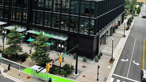 Un edificio moderno de gran altura con ventanas de vidrio reflectante, un carril bici verde y una acera con mesas al aire libre.