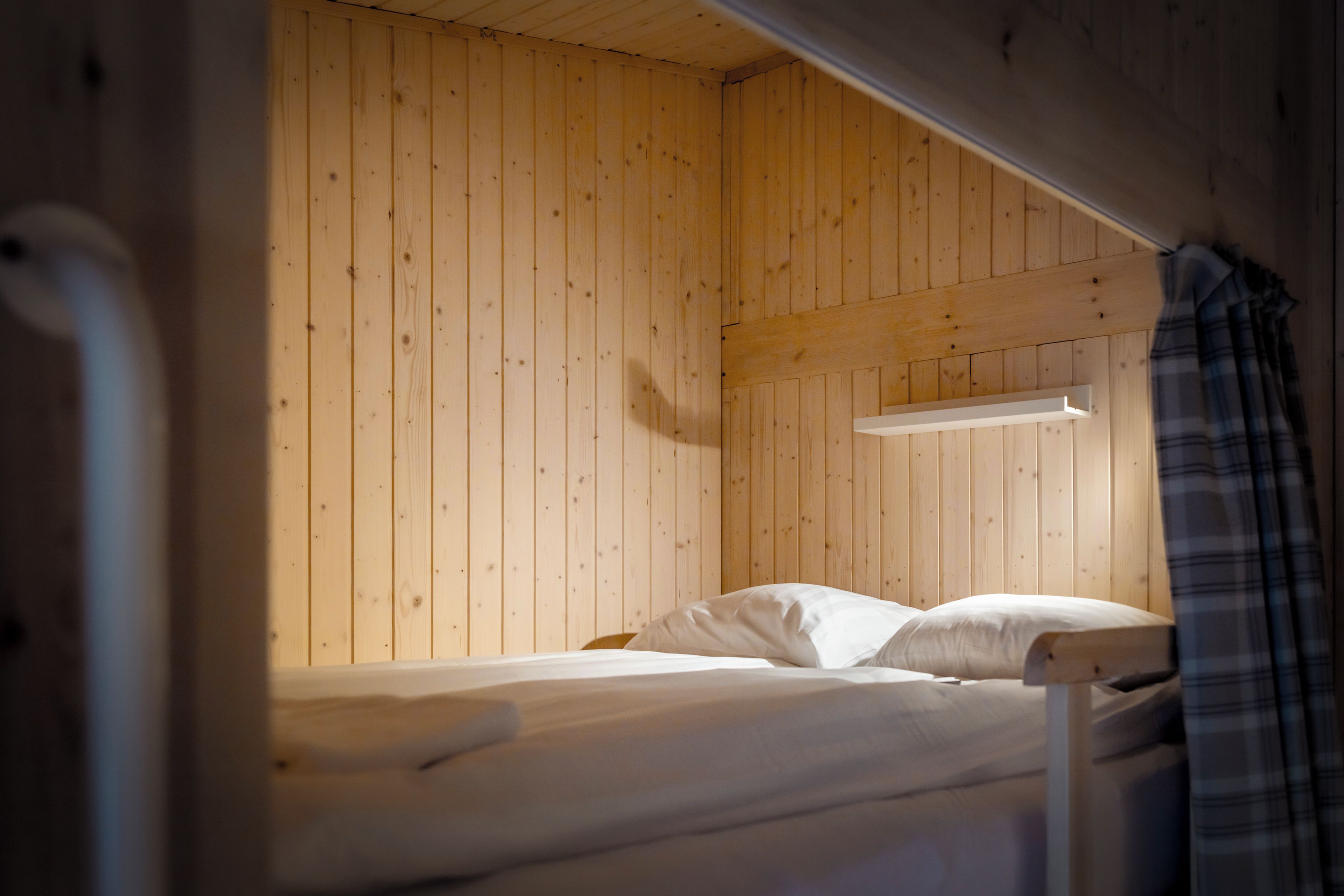Una cama tipo litera con un panel de madera, un estante pequeño y una cortina escocesa.