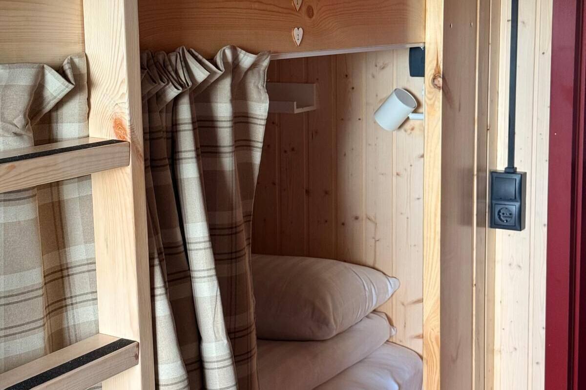 Ein hölzernes Etagenbett mit zwei Schlafstellen, jeweils mit Kissen und Decke. Auf der rechten Seite befindet sich ein Regal mit gefalteten Decken und einer kleinen weißen Tasse.