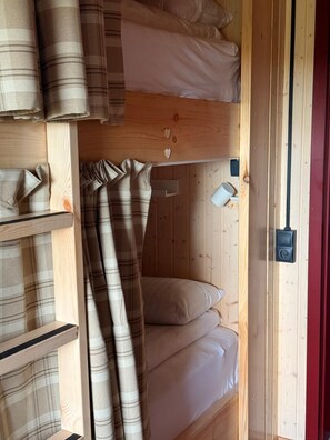 A wooden bunk bed with two levels, each with a pillow and a blanket. There is a shelf with folded blankets and a small white cup on the right side. - The Fox Hostel (Hrífunesvegur)