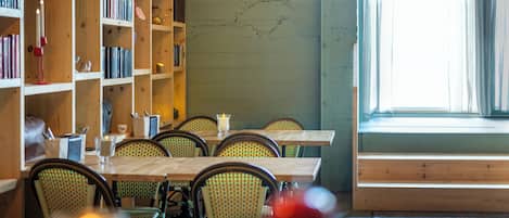 A dining area with wooden tables and chairs, a bookshelf with books, and a painting on the wall.