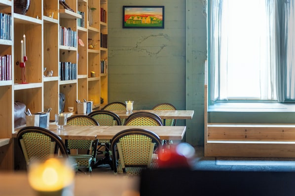 Un coin repas avec des tables et des chaises en bois, une bibliothèque remplie de livres et un tableau accroché au mur.