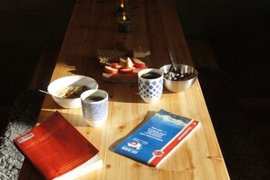A wooden table with a book, a cup of coffee, a bowl of cereal, a mug, and a plate of fruit. - The Fox Hostel (Hrífunesvegur)