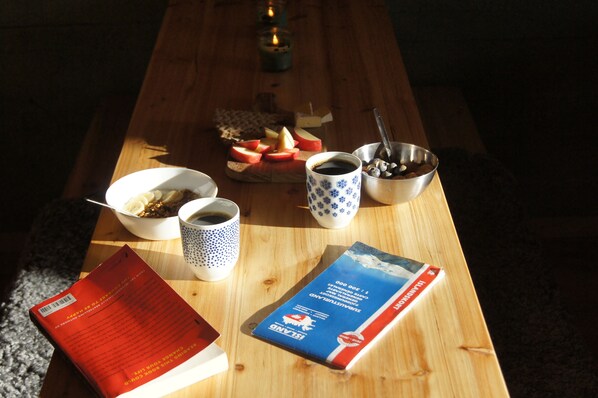 A wooden table with a book, a cup of coffee, a bowl of cereal, a mug, and a plate of fruit. - The Fox Hostel (Hrífunesvegur)