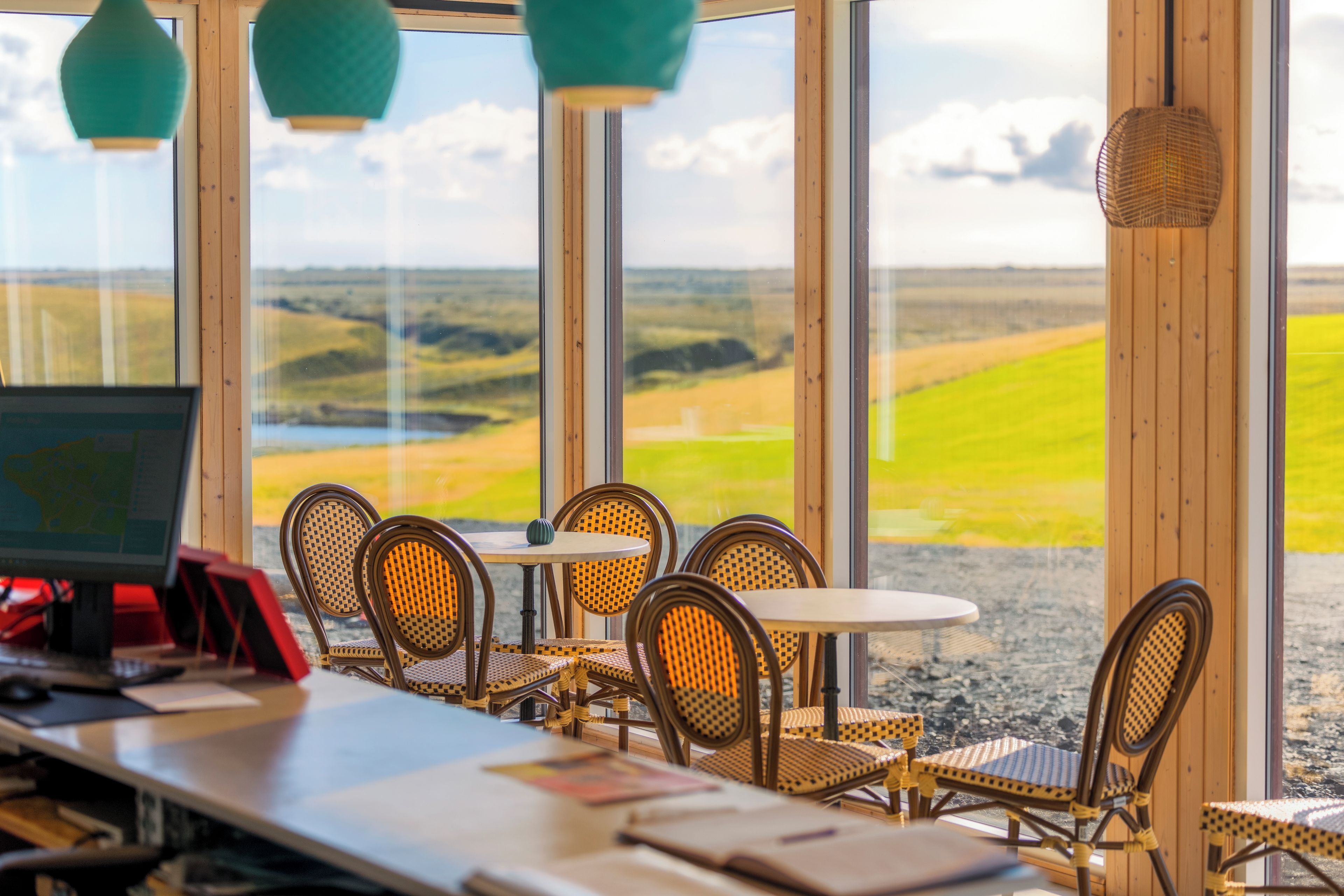 A dining area with wicker chairs and tables, a computer monitor, and a view of a landscape through large windows.