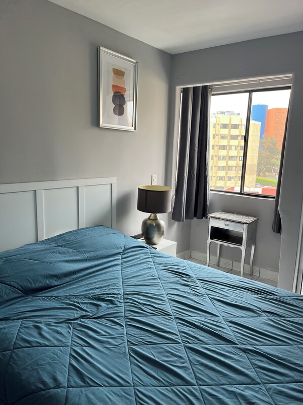 A bedroom with a bed, a bedside lamp, a small table, a window with curtains, and a framed picture on the wall.