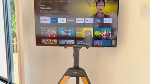 Un televisor montado sobre un trípode en una habitación con una pared lisa y una ventana.