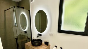 Un baño moderno con cabina de ducha de vidrio, tocador de madera con lavamanos negro y dos espejos redondos.