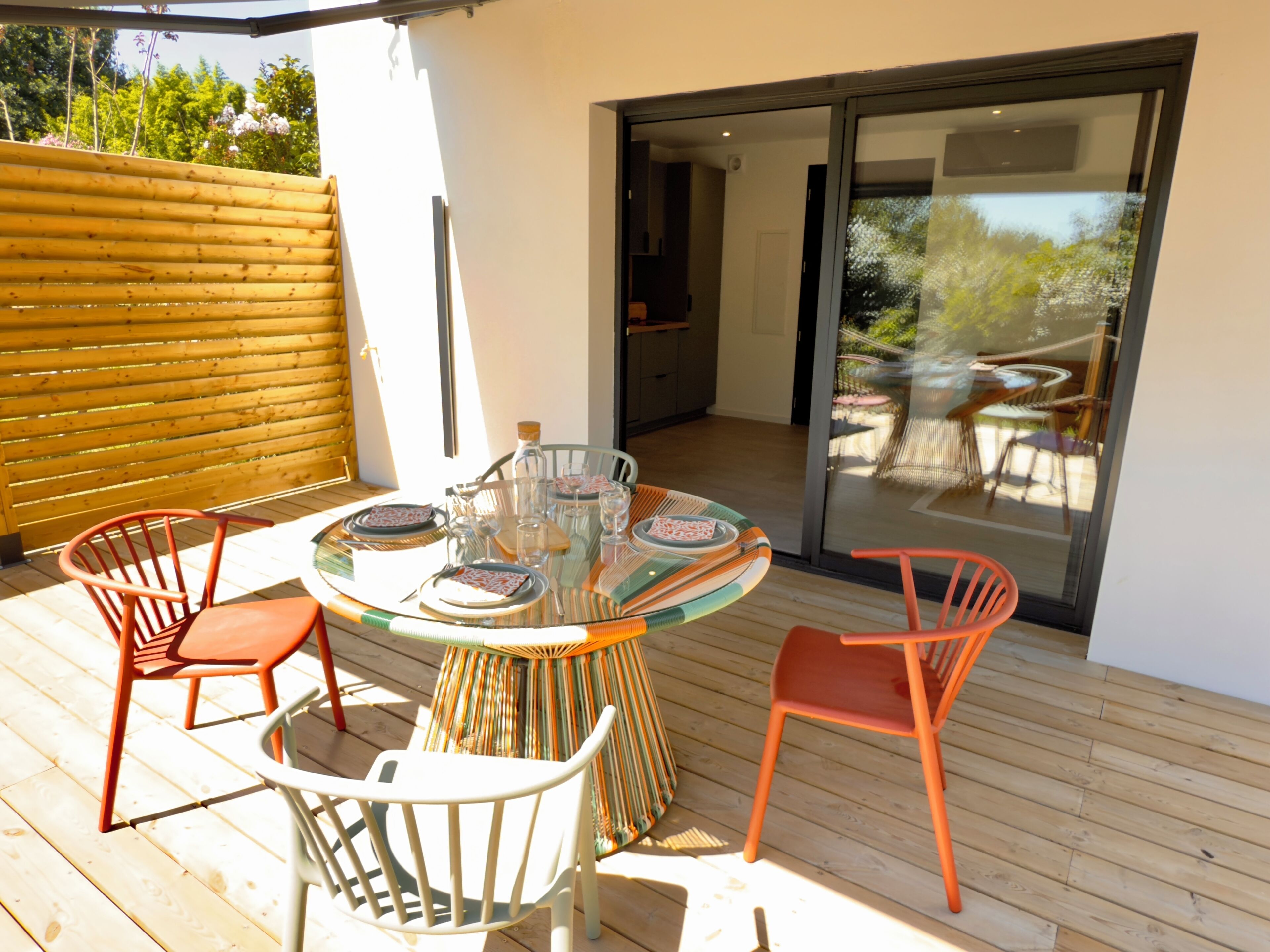 An outdoor dining area with a glass table surrounded by chairs, a wooden deck, and a sliding glass door leading to a patio.