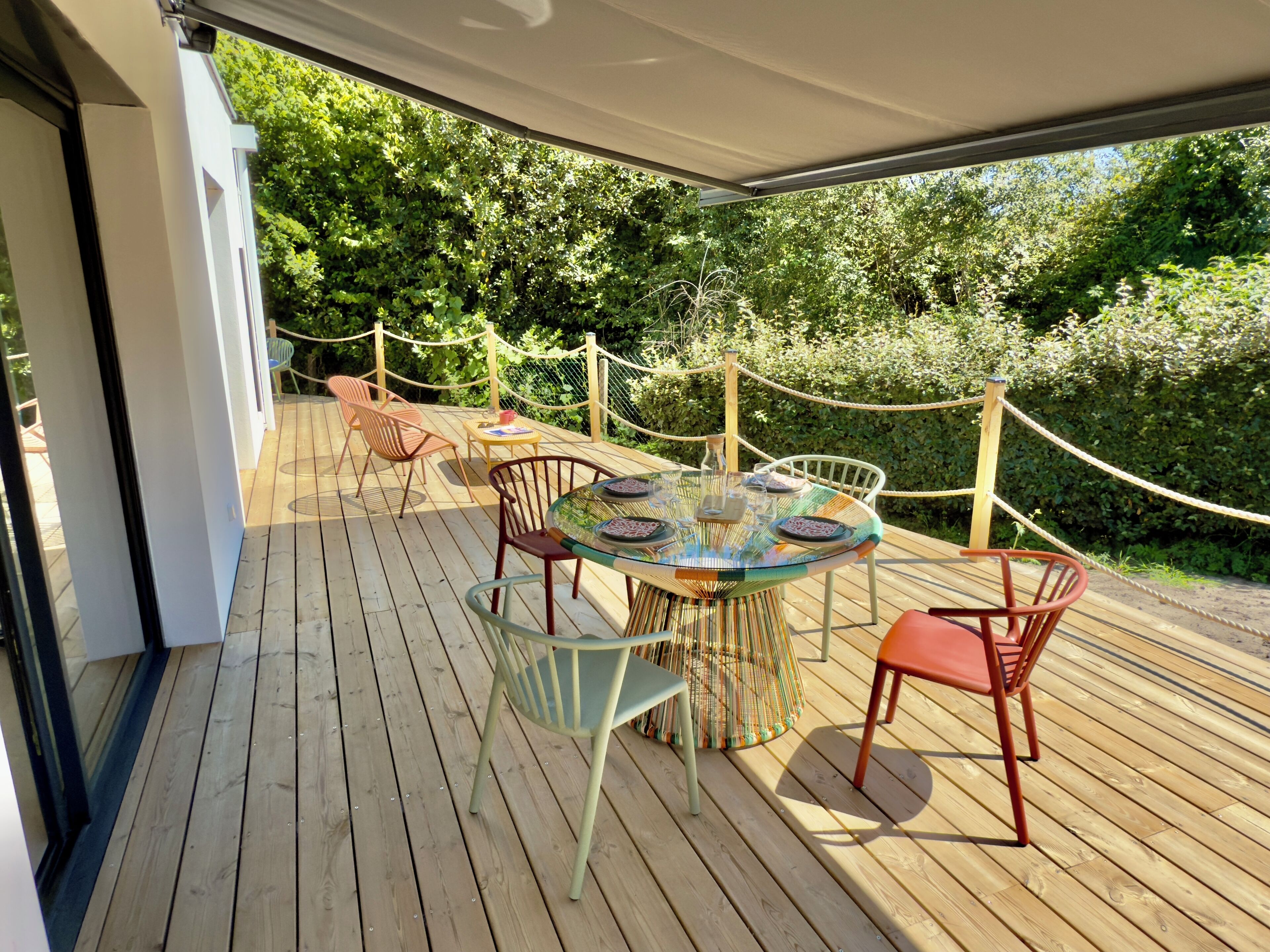 Une terrasse extérieure en bois, avec une table ronde et des chaises, entourée d’une barrière en corde et d’une végétation luxuriante.
