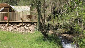 A rustic wooden cabin with a deck, surrounded by lush greenery and a small stream. - ZEcamping toboggans piscine animaux acceptés locatif 16m2 4 pers (Saint-Constant-Fournoulès)