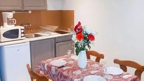 A compact kitchen with a microwave, coffee maker, and a small refrigerator. A dining table with a floral tablecloth and a vase of flowers is set for four.