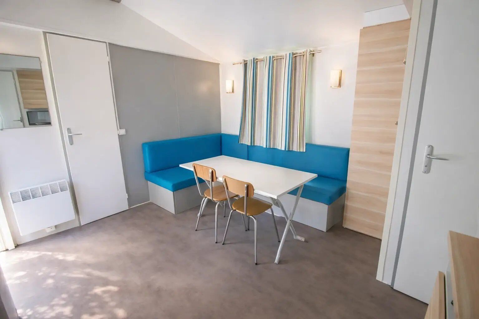 A compact room with a white table, two chairs, a blue bench, and a window with striped curtains.