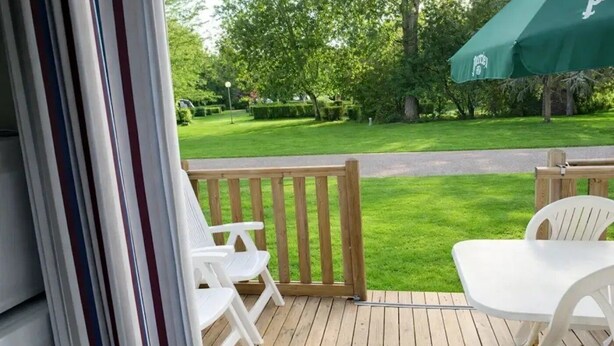 Een houten terras met witte plastic stoelen en een tafel, met uitzicht op een verzorgd gazon en bomen.