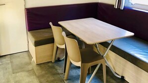 A compact room with a wooden table and chairs, a bench, a small cabinet, and a window with curtains. - ZEcamping 3 étoiles piscine chauffée mobil-home TV clim 33m2 6 pers (Portel-des-Corbières)