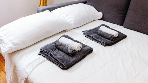 Una cama bien tendida con ropa de cama blanca y gris, dos toallas dobladas y una cortina amarilla.