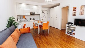 Una sala de estar moderna con un sofá gris, cojines naranja, una zona de comedor con una mesa blanca y sillas naranja, una cocina con armarios y electrodomésticos blancos, y un televisor sobre un mueble blanco.