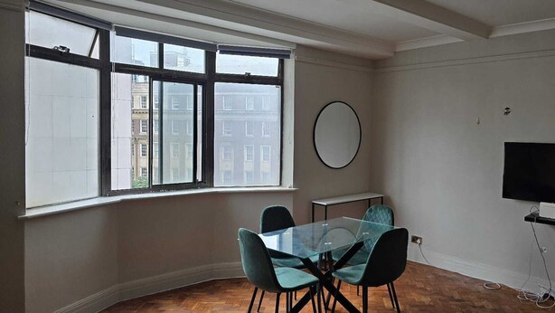 A room with a large window, a round mirror, a glass table with chairs, and a TV mounted on the wall.