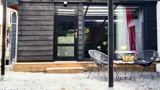 Un espacio al aire libre techado con estructura de madera, puerta corredera de vidrio, dos sillas de mimbre y una pequeña mesa.